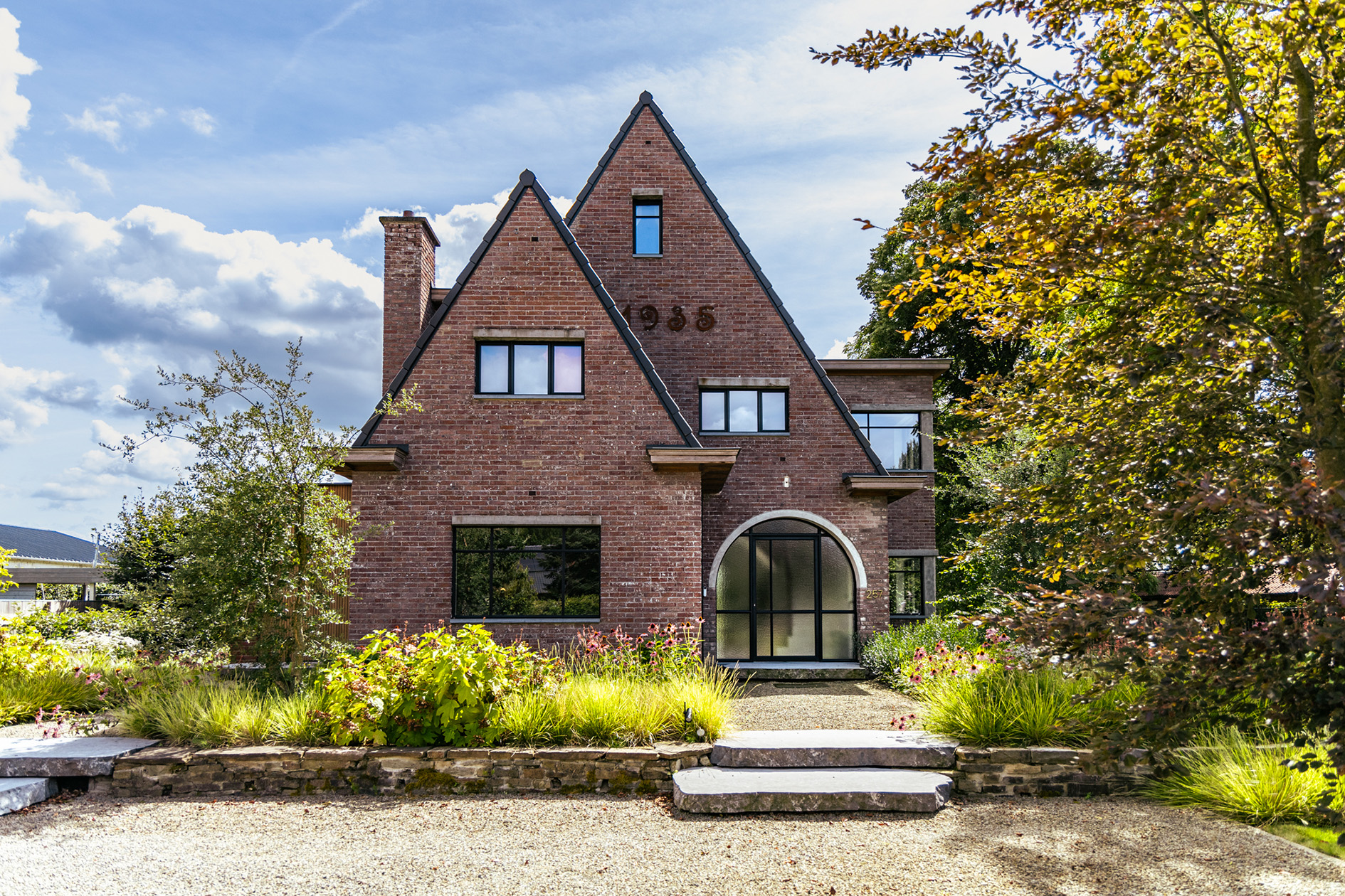 Bakstenen huis met twee steile puntdaken, zwarte ramen en een halve cirkelvormige glazen deur, omringd door groen en struiken.