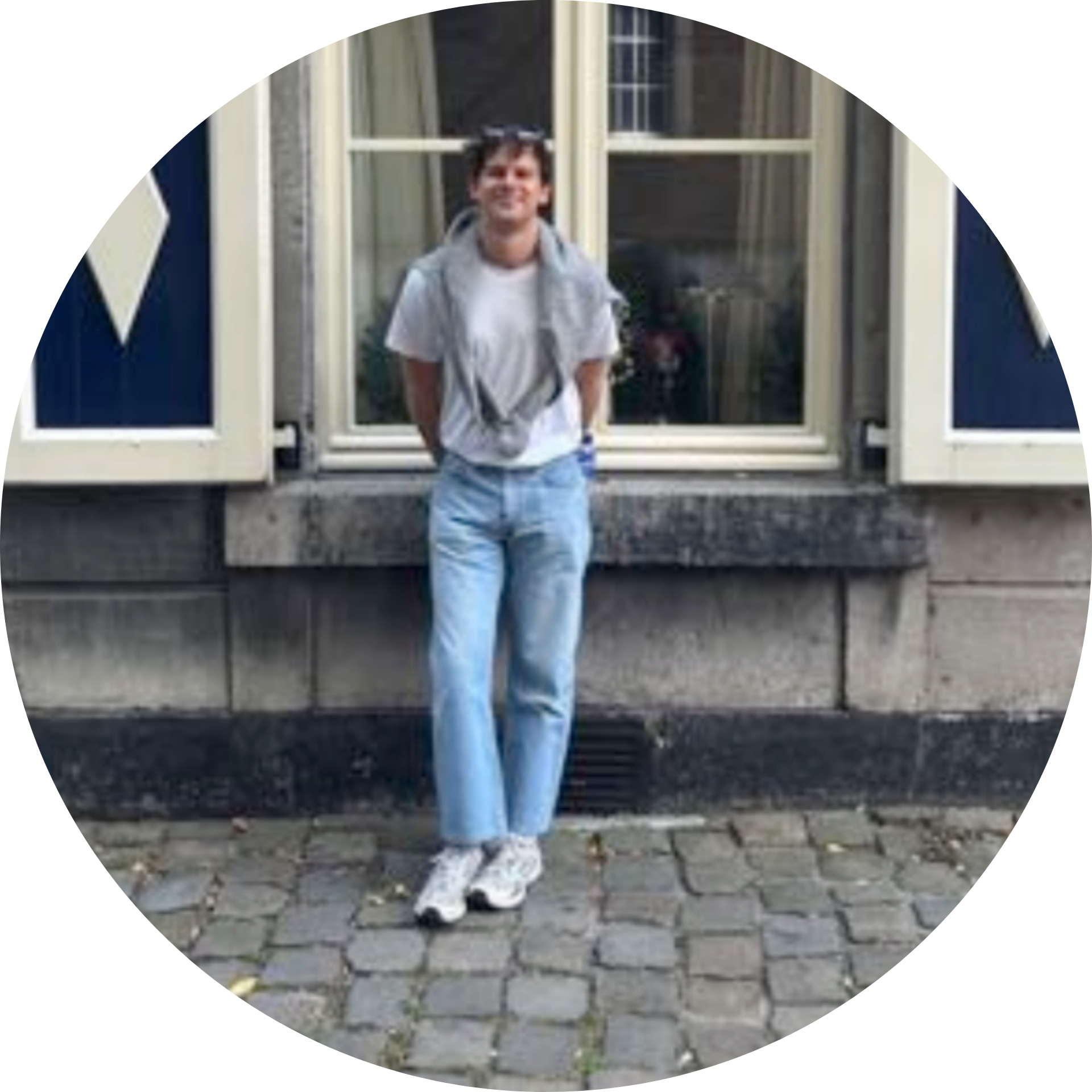 Smiling young man wearing light blue jeans, white t-shirt, and gray sweater draped over shoulders, standing on a cobblestone street in front of a window with cream-colored shutters.